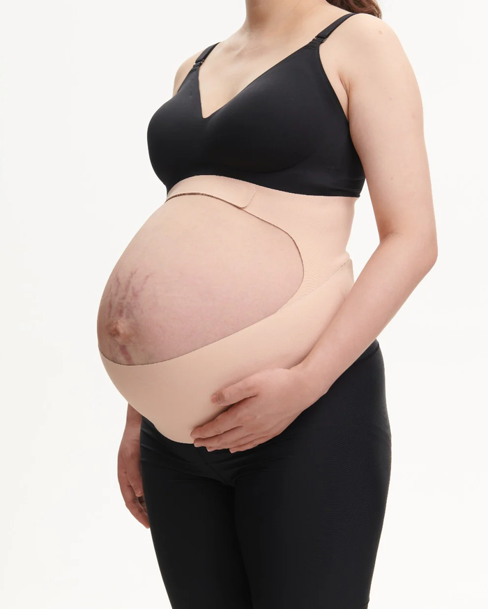 Pregnant person modeling a beige BumpEase Momcozy Ergonest maternity belly band over black leggings and nursing bra.