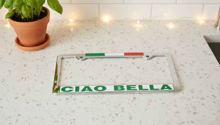 License plate frame with 'CIAO BELLA' text on a countertop with a plant and lights in the background