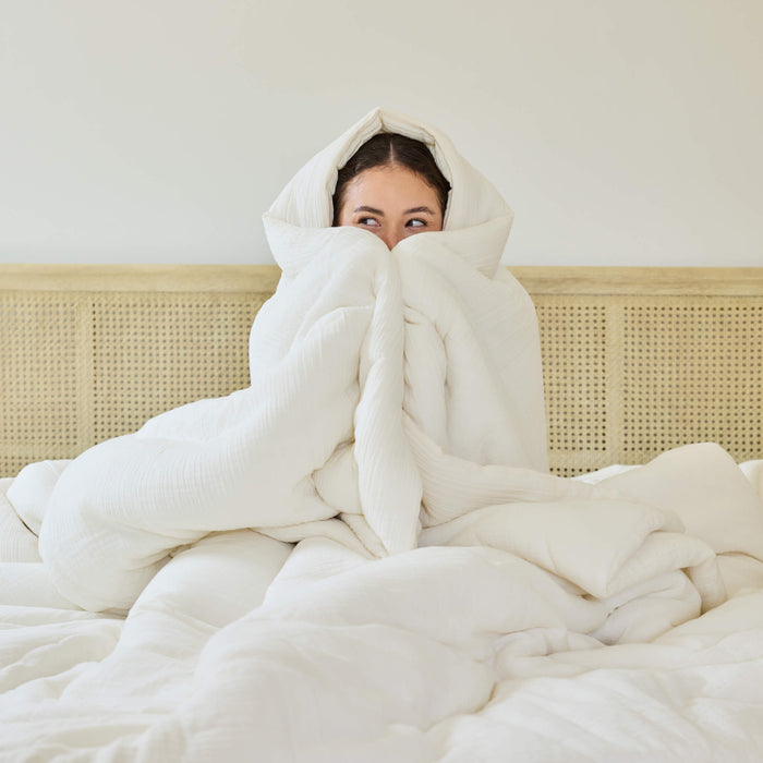 A young woman wrapped in an Avec Goods Cotton Quilt while sitting on a bed