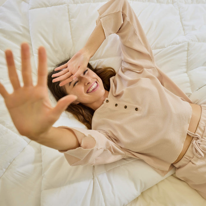 A young woman lying on a bed made with Avec Goods bedding while holding her hand up to the camera and smiling