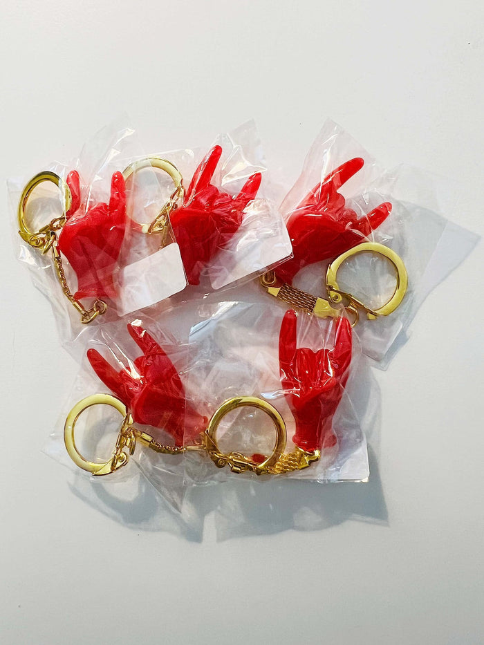 Red hand-shaped keychain making the “rock on” gesture, attached to a gold-colored chain and keyring in pack of 5. The glossy charm blends Italian Malocchio symbolism with rock culture, serving as a bold accessory for protection, style, or gifting.