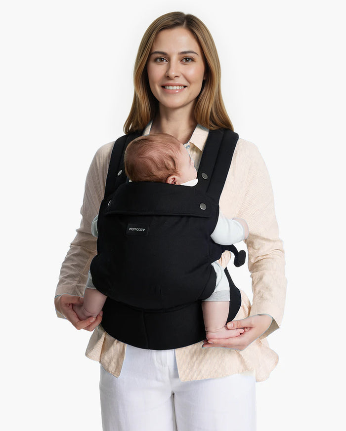 Momcozy PureHug Baby Carrier. Black carrier, comfortable design, worn by mother holding baby. Ideal for babywearing.