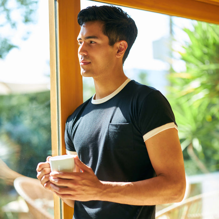 A young man holding a cup of coffee while looking out the window and wearing a Slumber Cloud UltraCool Pocket Tee
